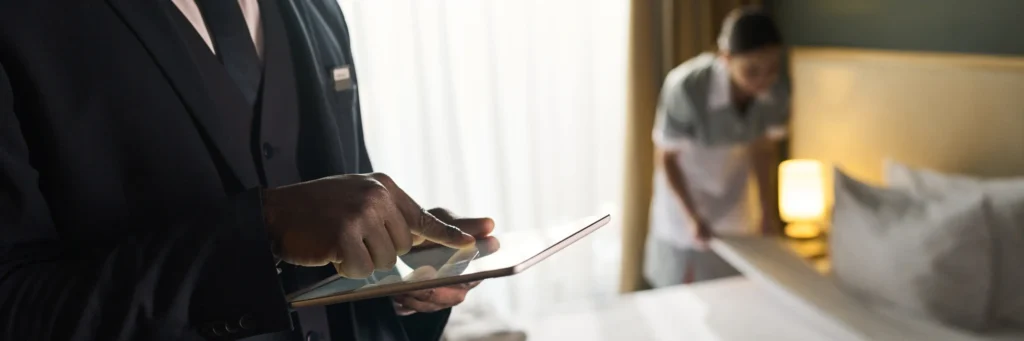Hotel manager using a tablet to improve digital booking strategies while housekeeping staff prepares a room