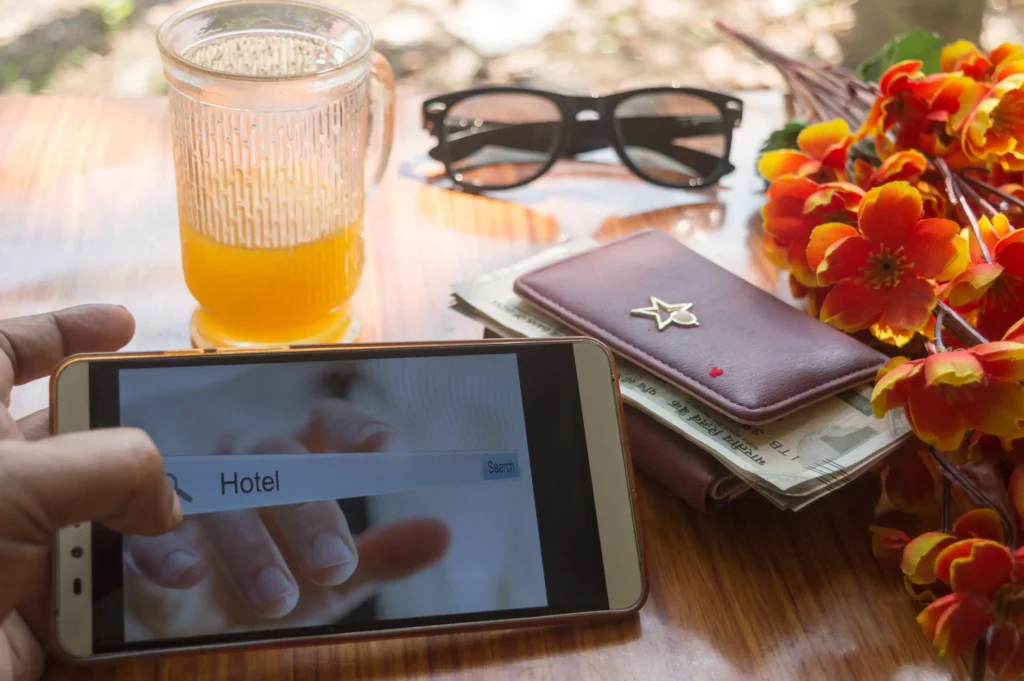 Traveler using mobile phone to search for hotel with drink, wallet, sunglasses and flowers on table